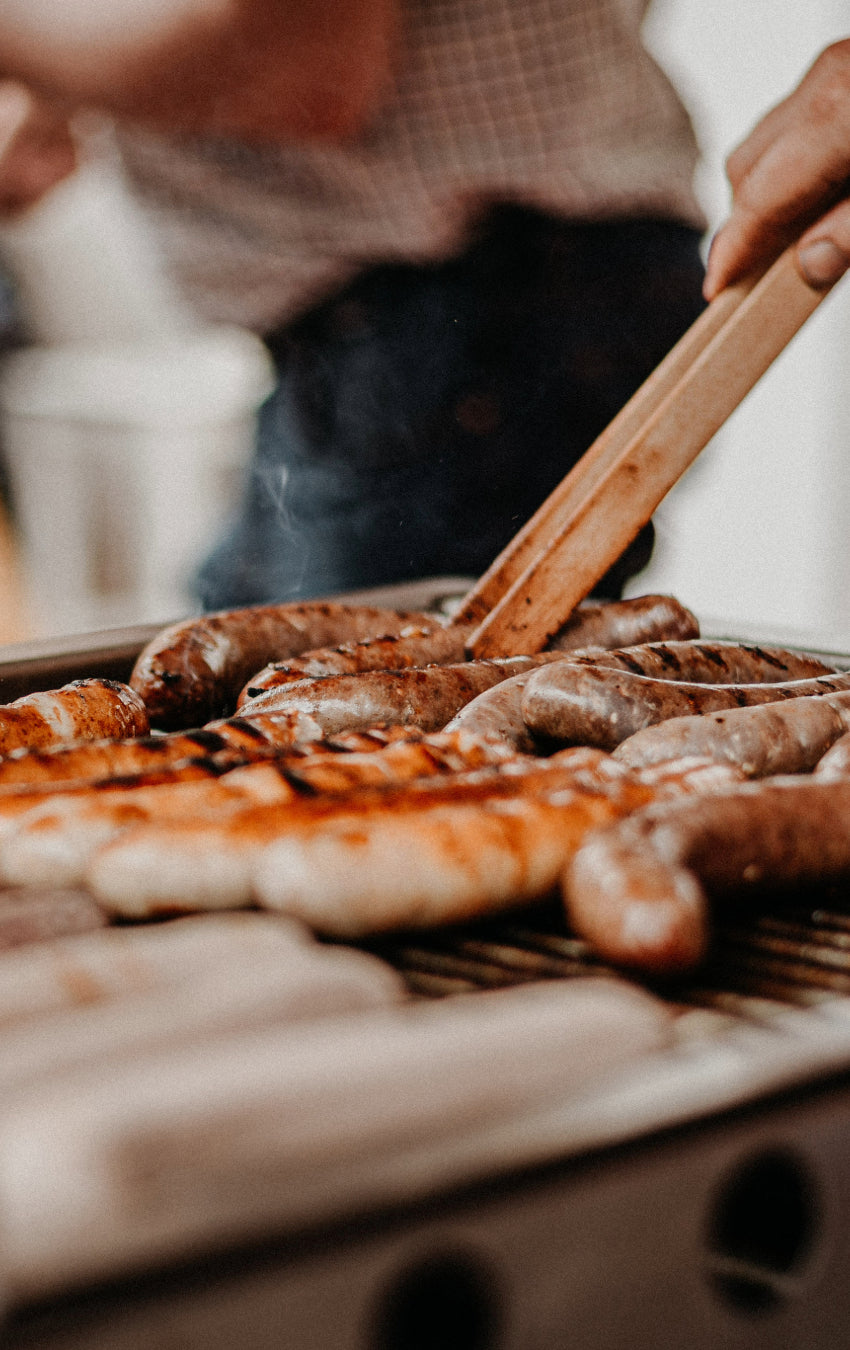Verschiedene Grill-Würste liegen auf dem Grill und eine Hand nimmt sich eine Grill-Wurst mit einer Grillzange.